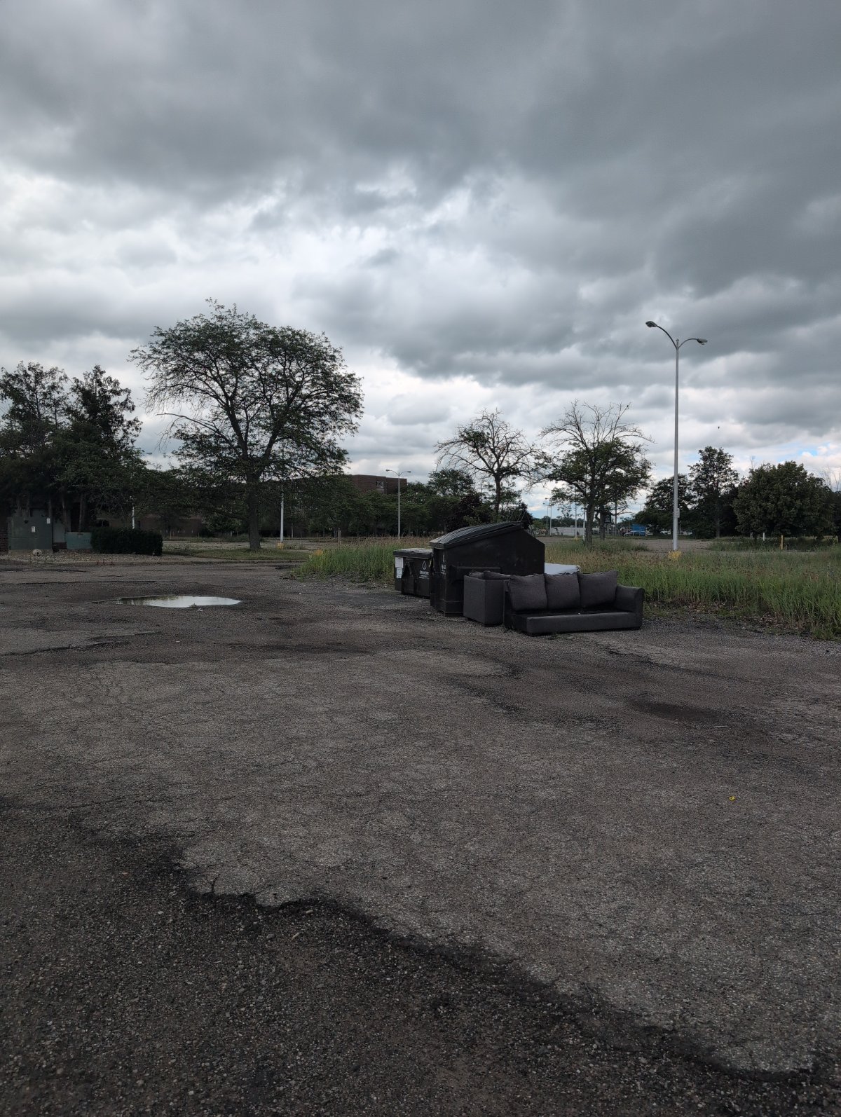 A discarded upholstered couch sitting alone in an open gravel lot under a dramatic, layered cloudy sky, with a bare tree and a lone lamppost on the horizon