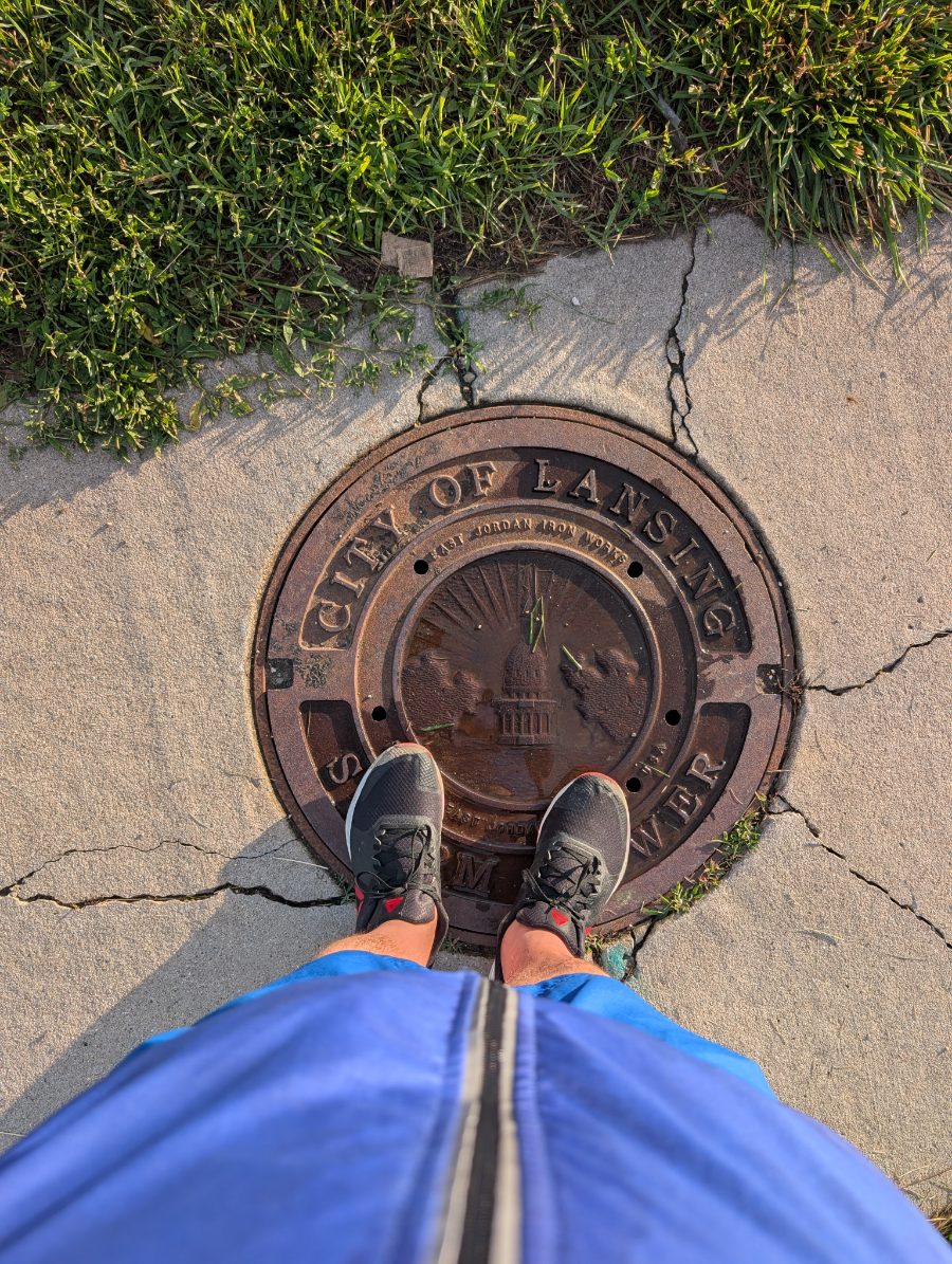 Album cover for On God: the curator's own sneakers planted on a City of Lansing manhole cover (Liberty and a globe cast in the iron), cracked sidewalk and grass around — generic kicks, standing on the world.