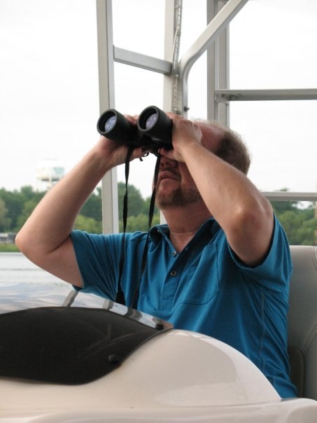 A man in a teal polo shirt on a pontoon boat, looking through binoculars out over the water