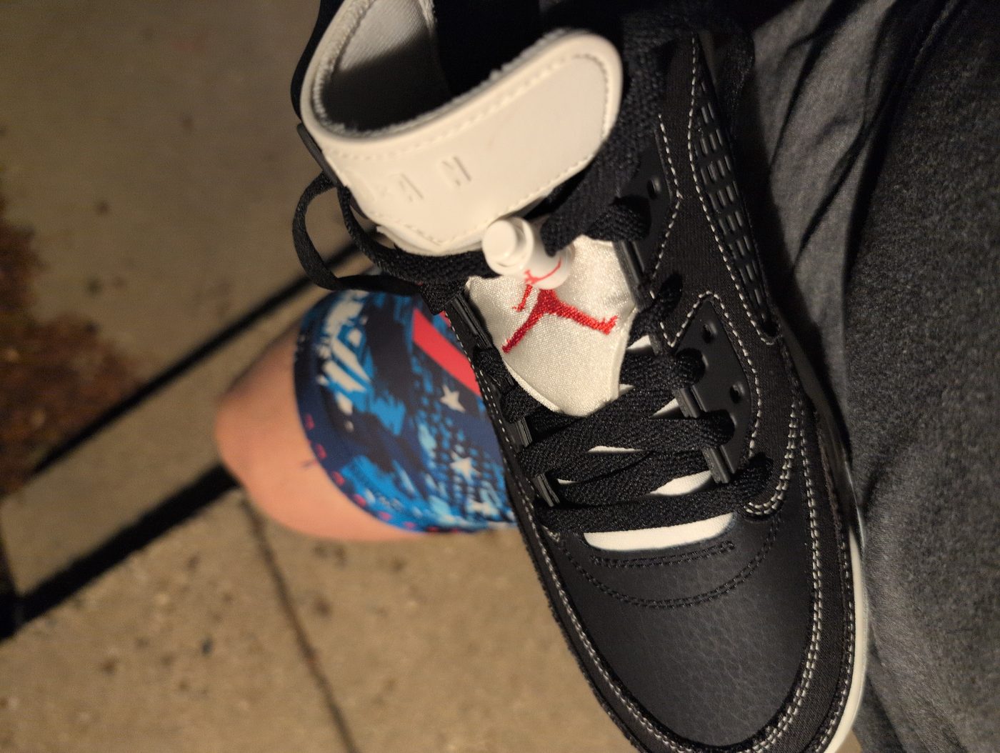 Black Air Jordans with a red Jumpman on the tongue, worn with gray sweatpants and stars-and-stripes socks, on a sidewalk at night