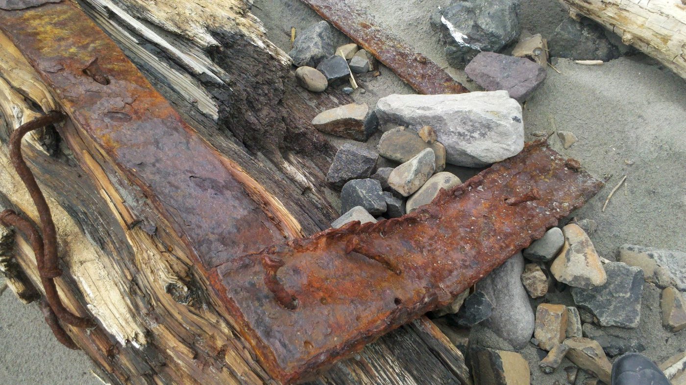 A heavily rusted metal door frame lying among weathered driftwood and smooth beach stones on sand