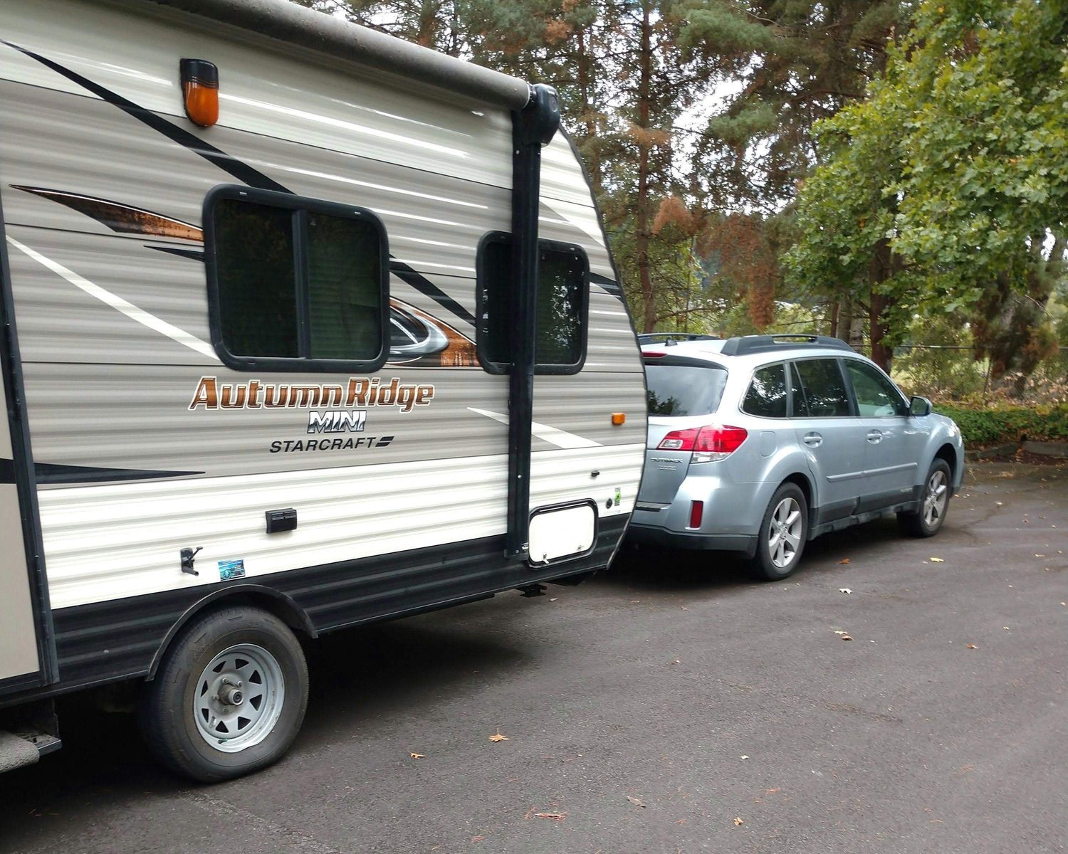 A silver Subaru Outback hitched to a Starcraft Autumn Ridge Mini travel trailer, parked on pavement under tall pines and autumn trees