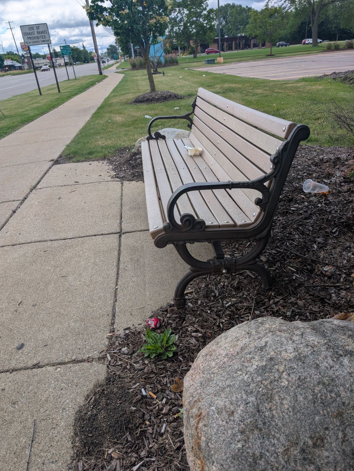 A park bench beside a busy road with litter and an empty parking lot behind it