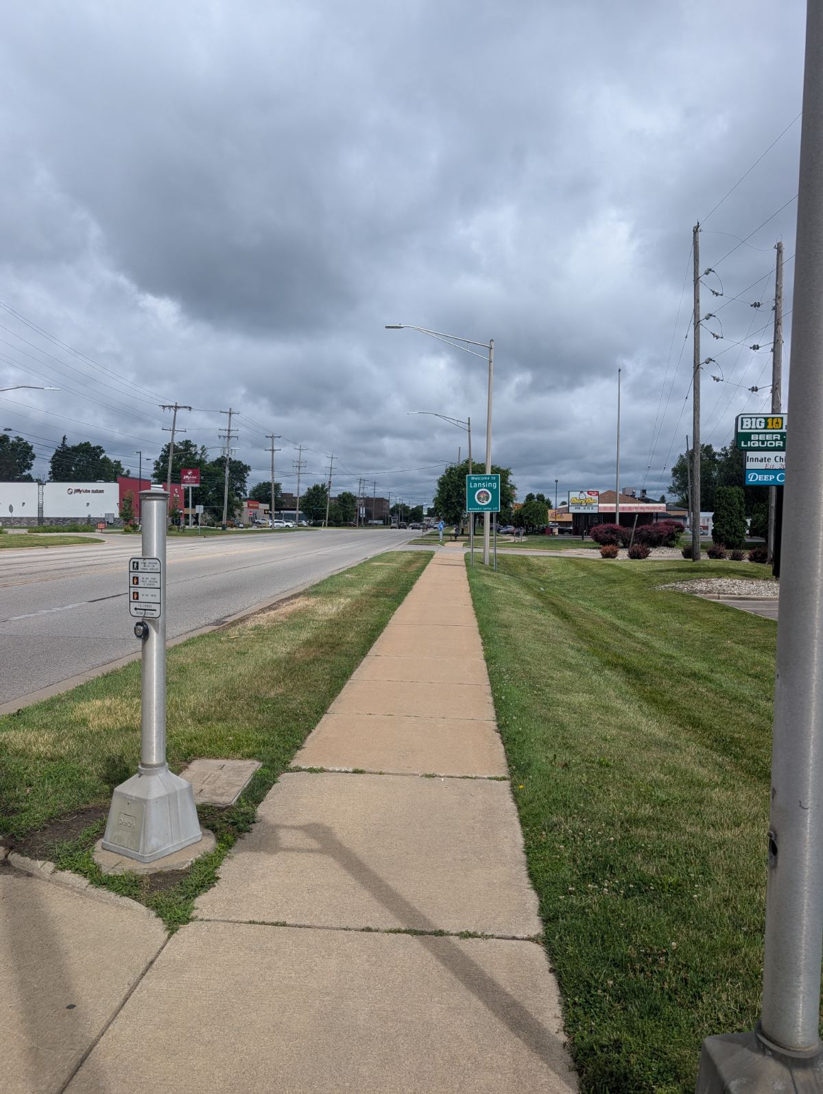 A Welcome to Lansing sign beside a sidewalk along a stroad under gray skies