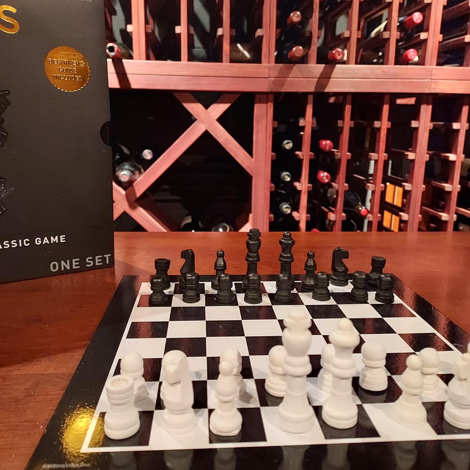 A chess set mid-setup on a wooden table in front of a full wine rack