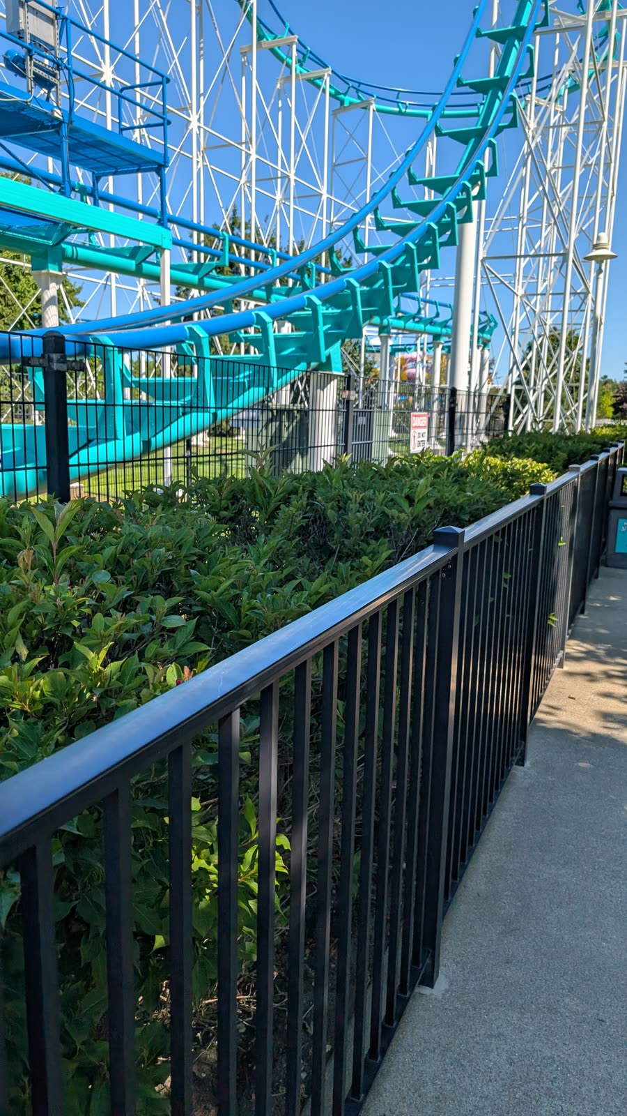 Teal and blue roller coaster track curving overhead above a park path and hedges
