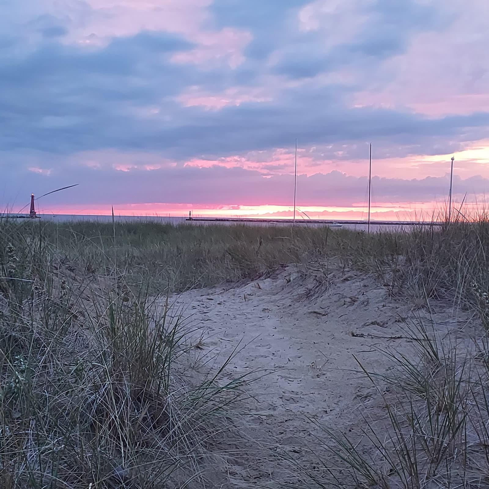 A Lake Michigan pier lighthouse and catwalk against a pink and purple sunset, dune grass in the foreground