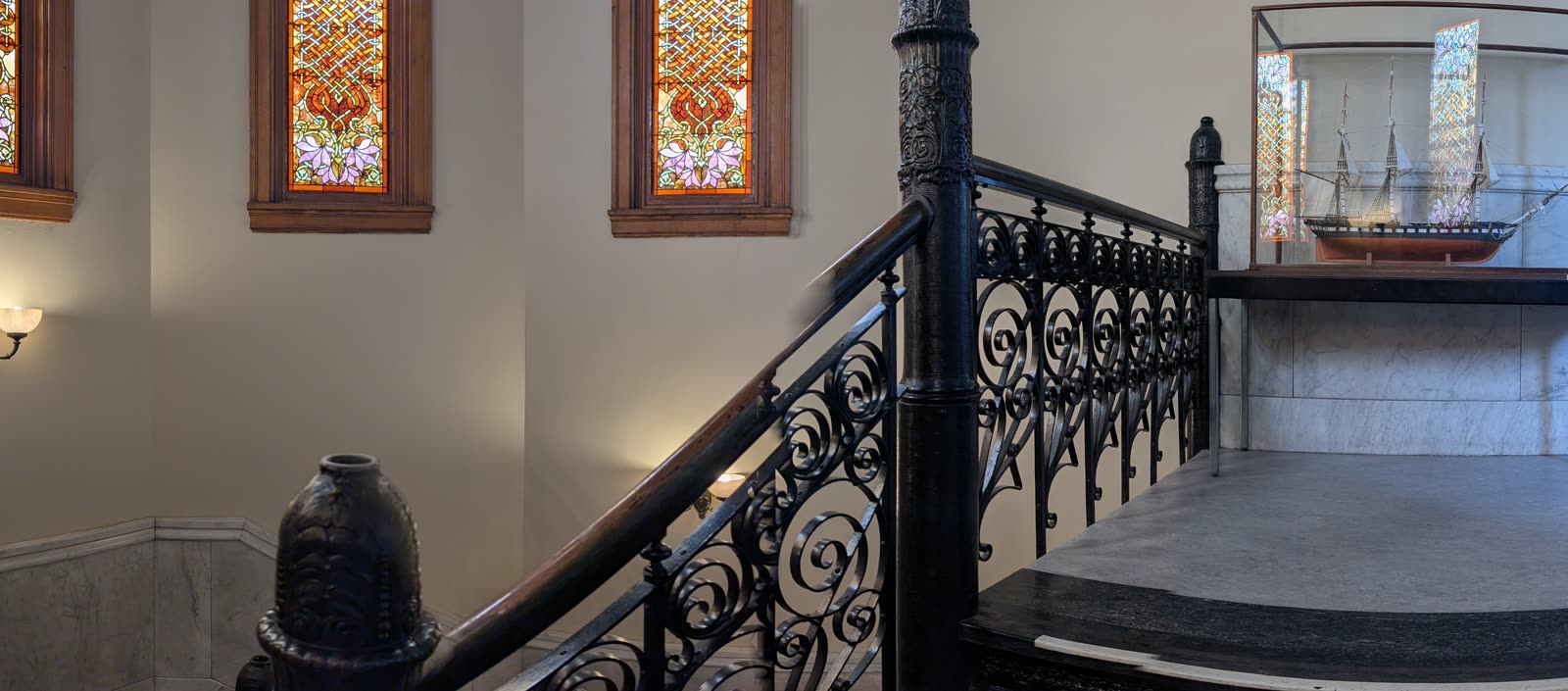 An ornate cast-iron staircase with stained-glass windows and a model ship in a case in a historic hall