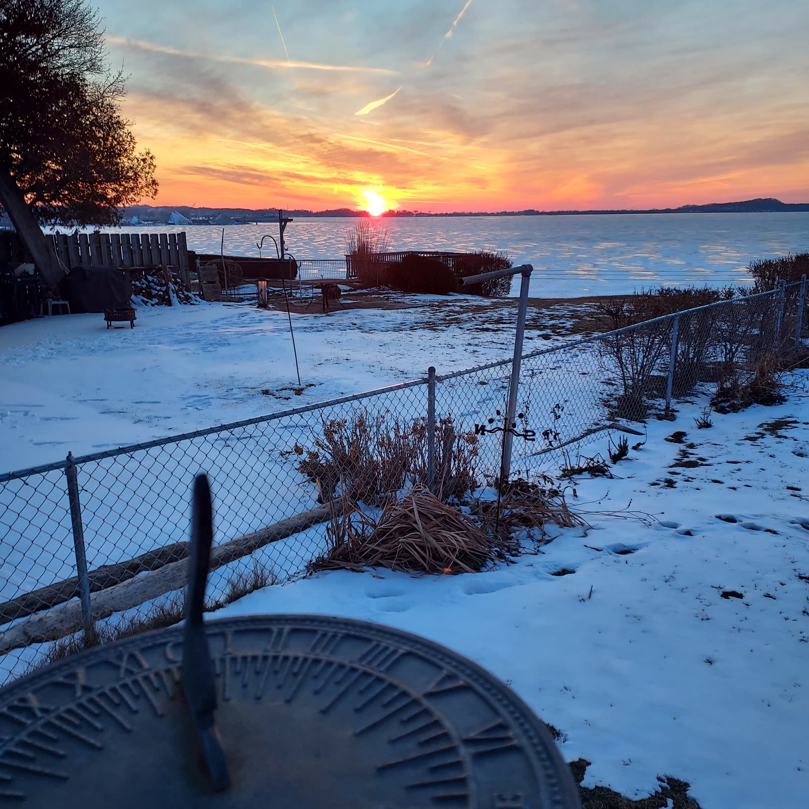 A sundial in the foreground with the real sun setting behind it over a frozen lake in snow