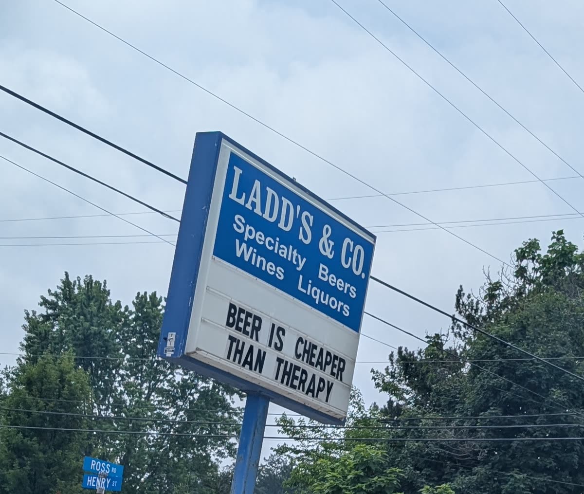A liquor-store marquee reading LADD'S and CO, Specialty Beers Wines Liquors, and BEER IS CHEAPER THAN THERAPY against a cloudy sky