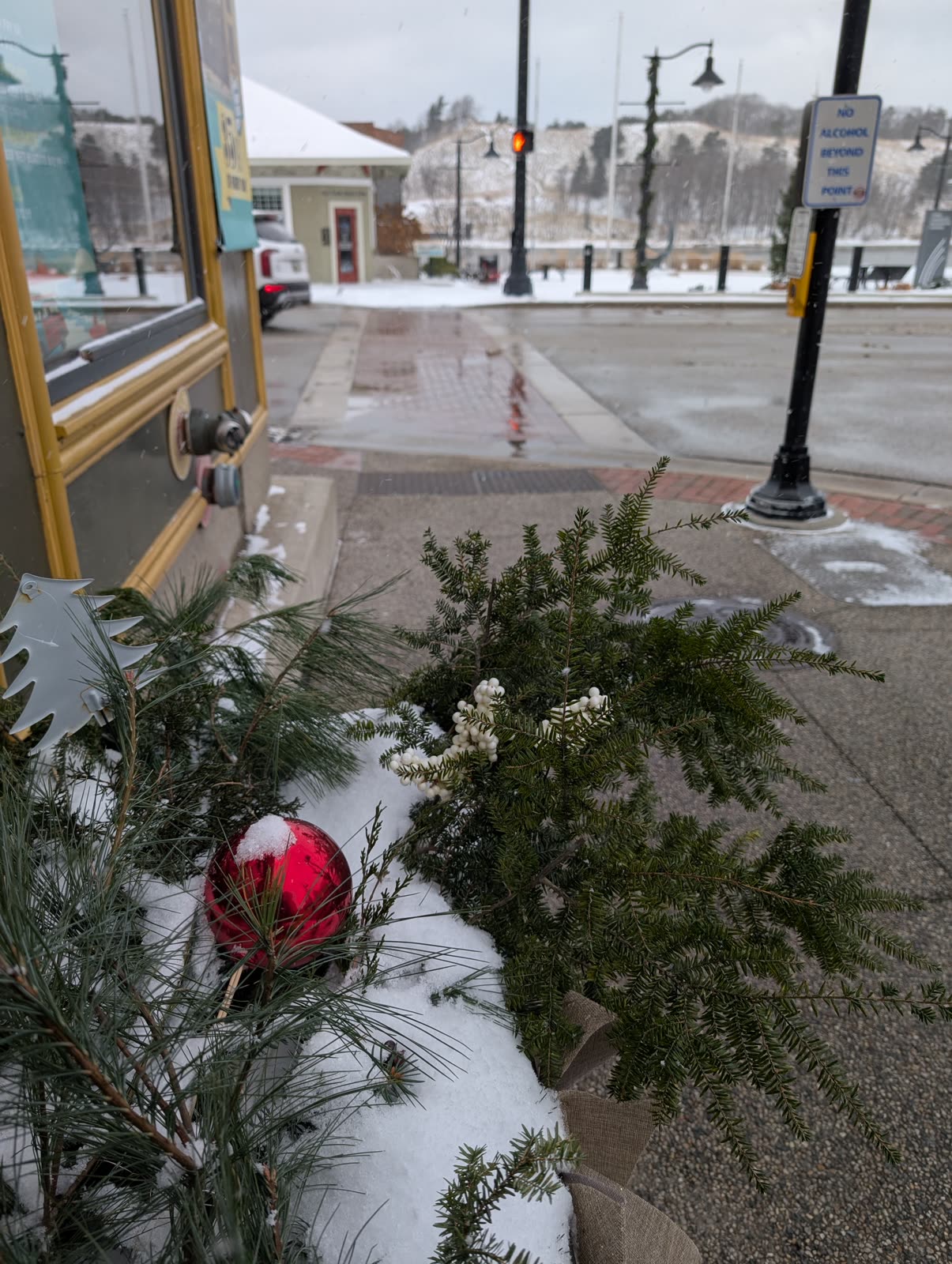 A snowy lakeside main street with holiday greenery, a red ornament, and a No Alcohol Beyond This Point sign